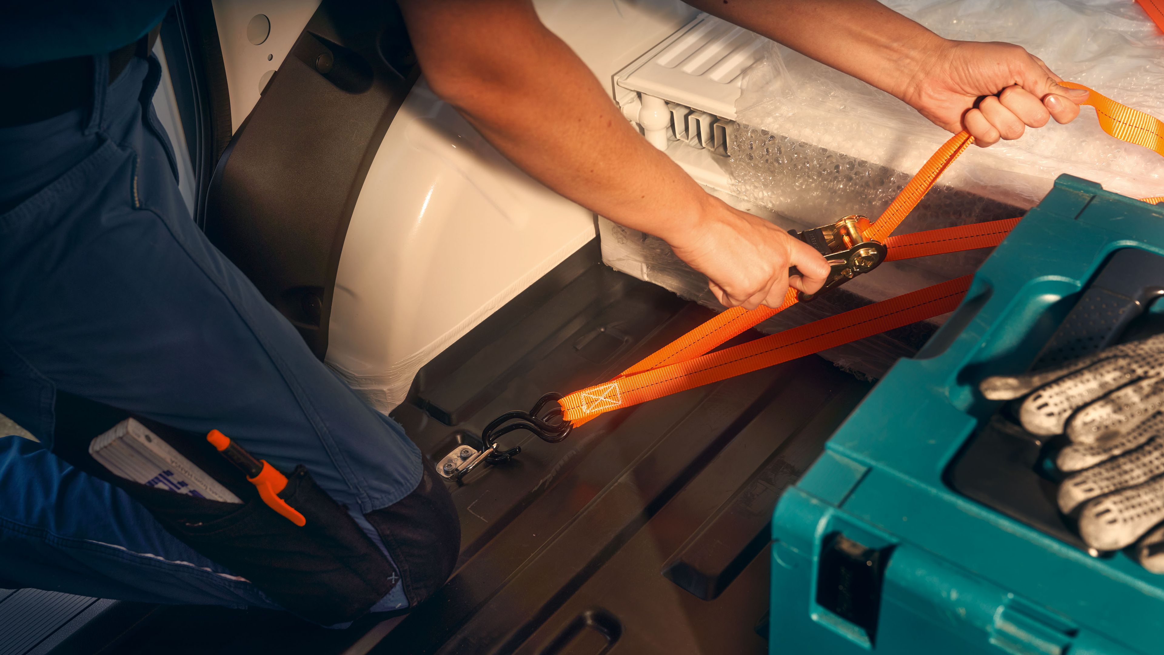 Pracovník uťahuje oranžový popruh okolo veľkého predmetu v zadnej časti vozidla. Vedľa neho na podlahe je zelená krabica s nástrojmi. Svetlo dopadá na ruky a predmet.