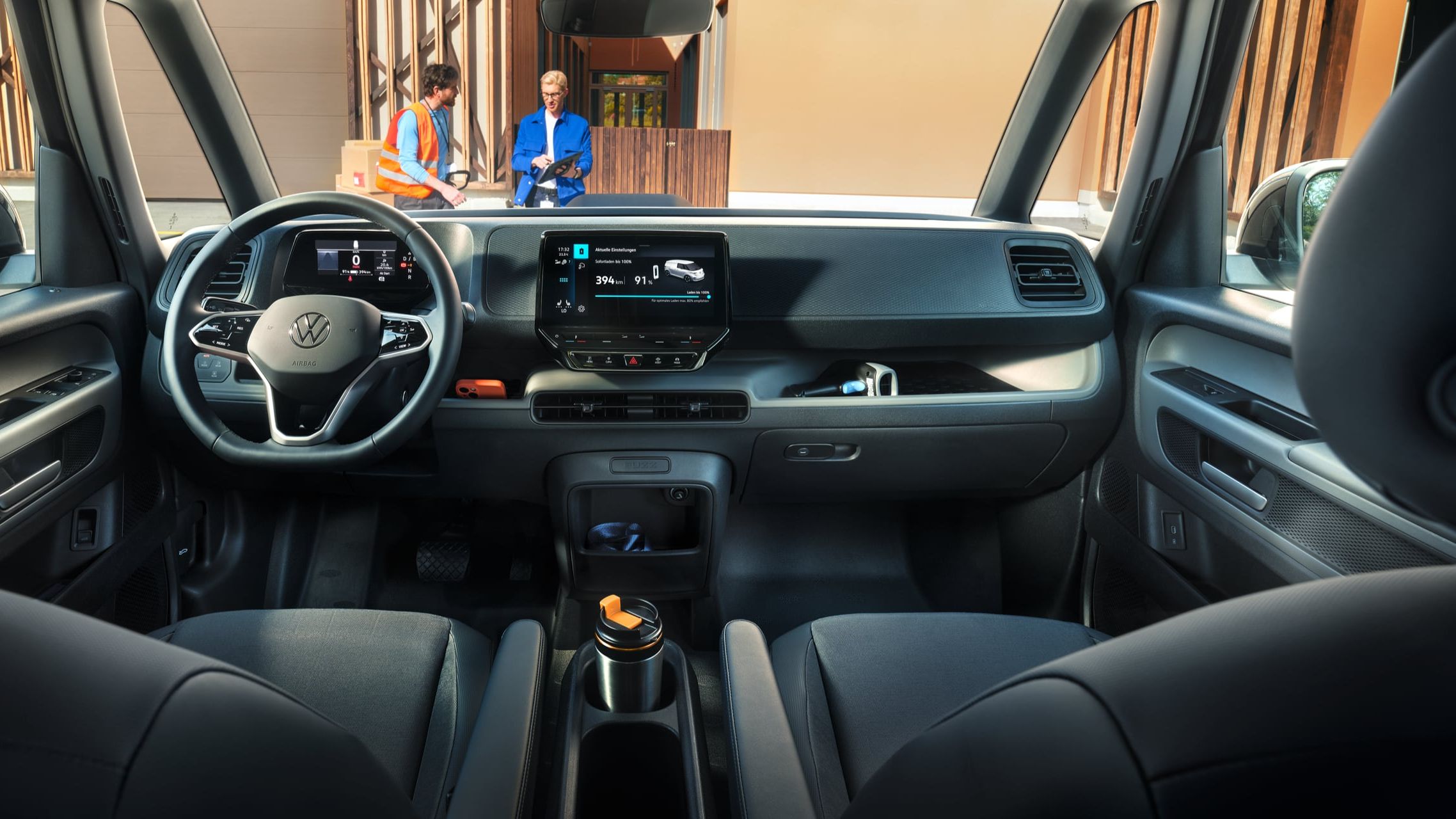 Pohľad do interiéru moderného VW ID. Buzz Cargo. Vidieť predné sedadlá, volant, prístrojový panel, palubná doska, stredová konzola s dotykovým displejom. Cez čelné okno vidieť dvoch ľudí, ktorí komunikujú pred budovou.