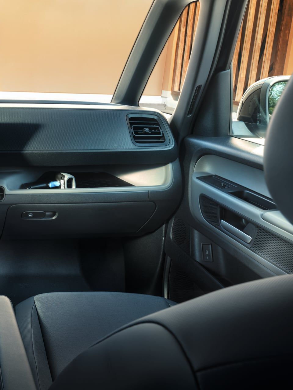 Pohľad do interiéru moderného VW ID. Buzz Cargo. Vidieť predné sedadlá, volant, prístrojový panel, palubná doska, stredová konzola s dotykovým displejom. Cez čelné okno vidieť dvoch ľudí, ktorí komunikujú pred budovou.