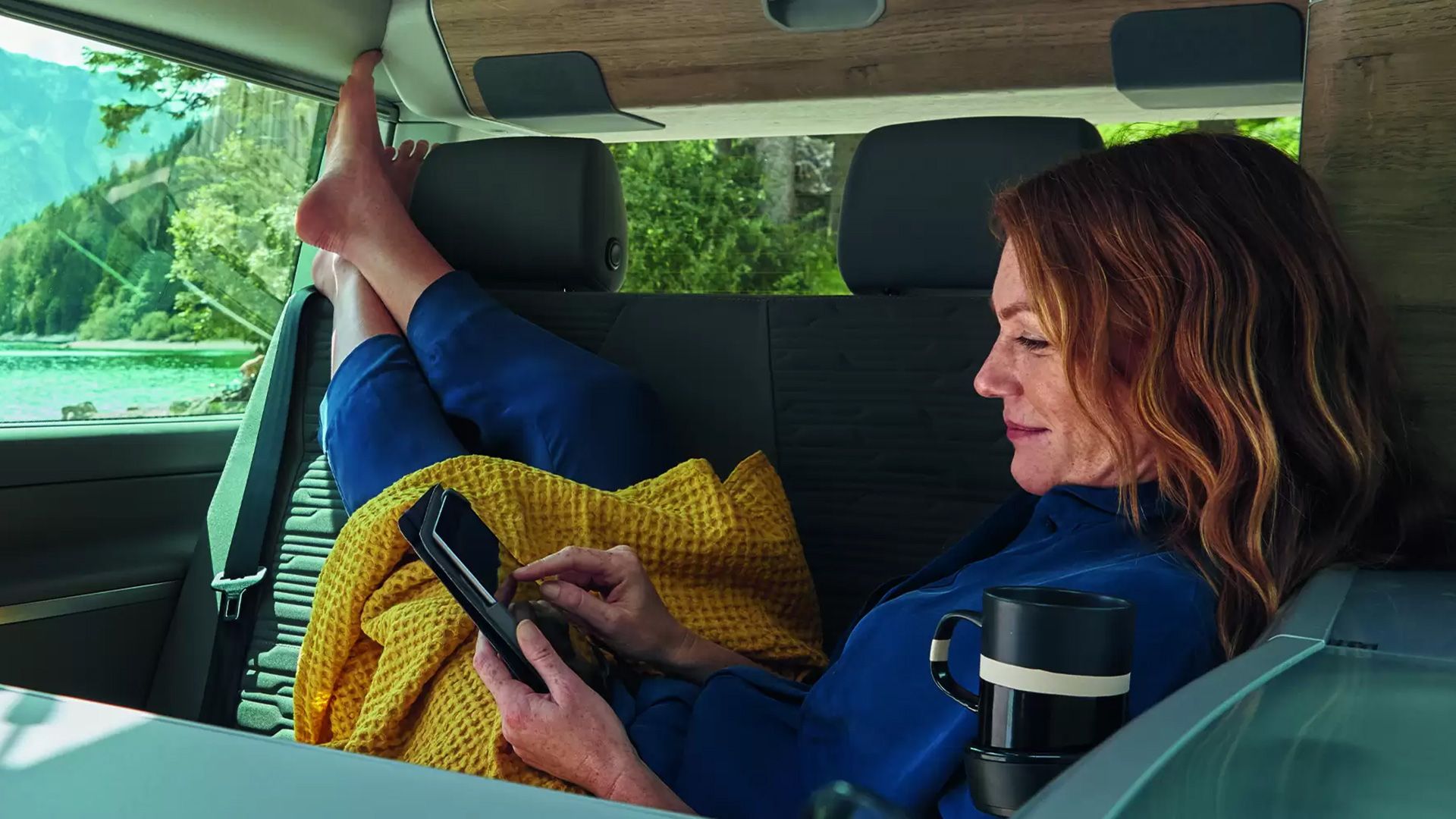 Žena sedí v aute, je oblečená v modrom oblečení, má nohy vyložené na sedadle a drží mobilný telefón. Vedľa nej sa nachádza šálka kávy, žena je zakrytá žltou dekou. V pozadí je výhľad na jazero, les, hory.