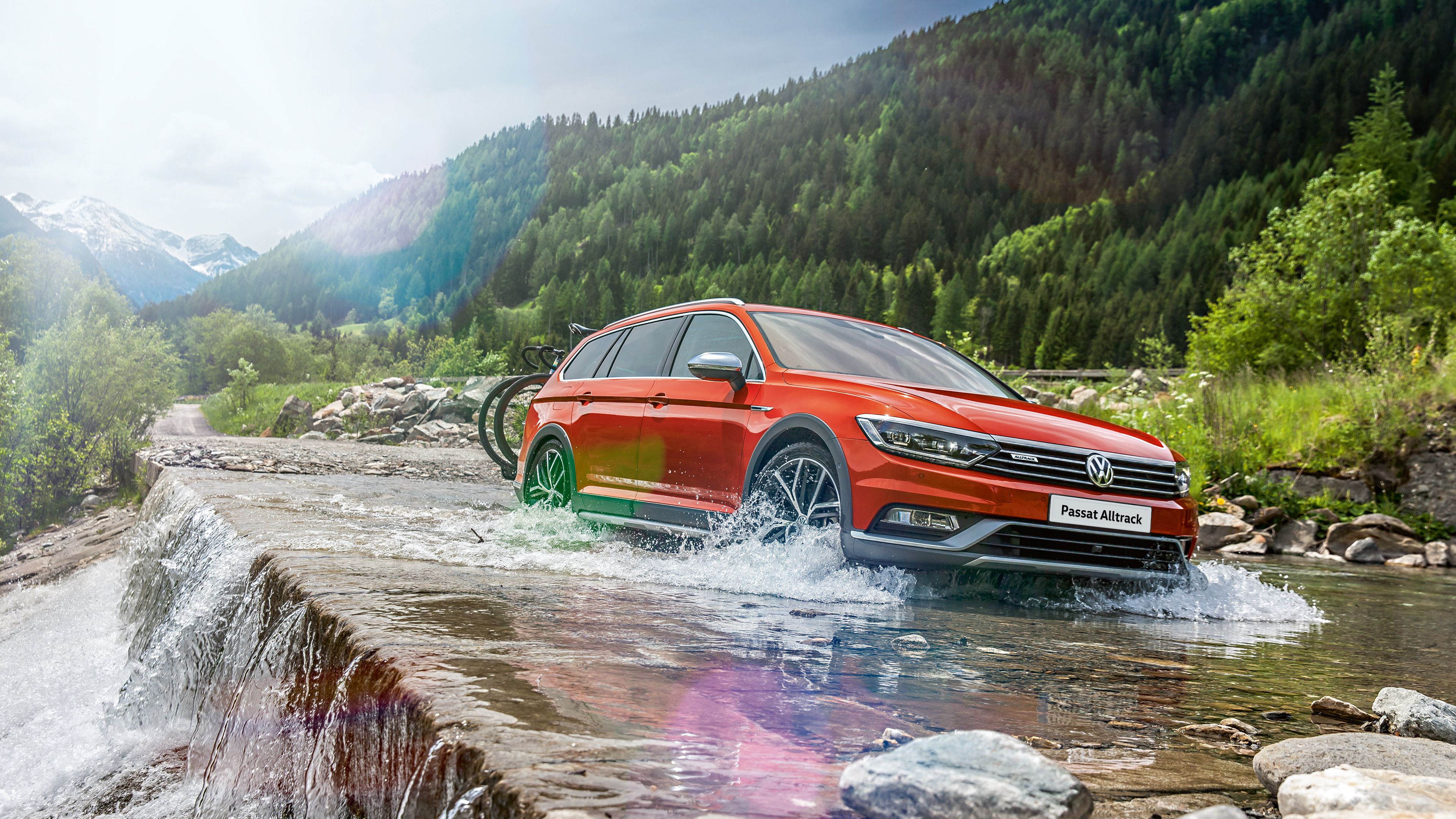 Červené auto Volkswagen Passat Alltrack prechádza cez plytký potok v horskom prostredí. V pozadí sú husté lesy a zasnežené vrcholky hôr.