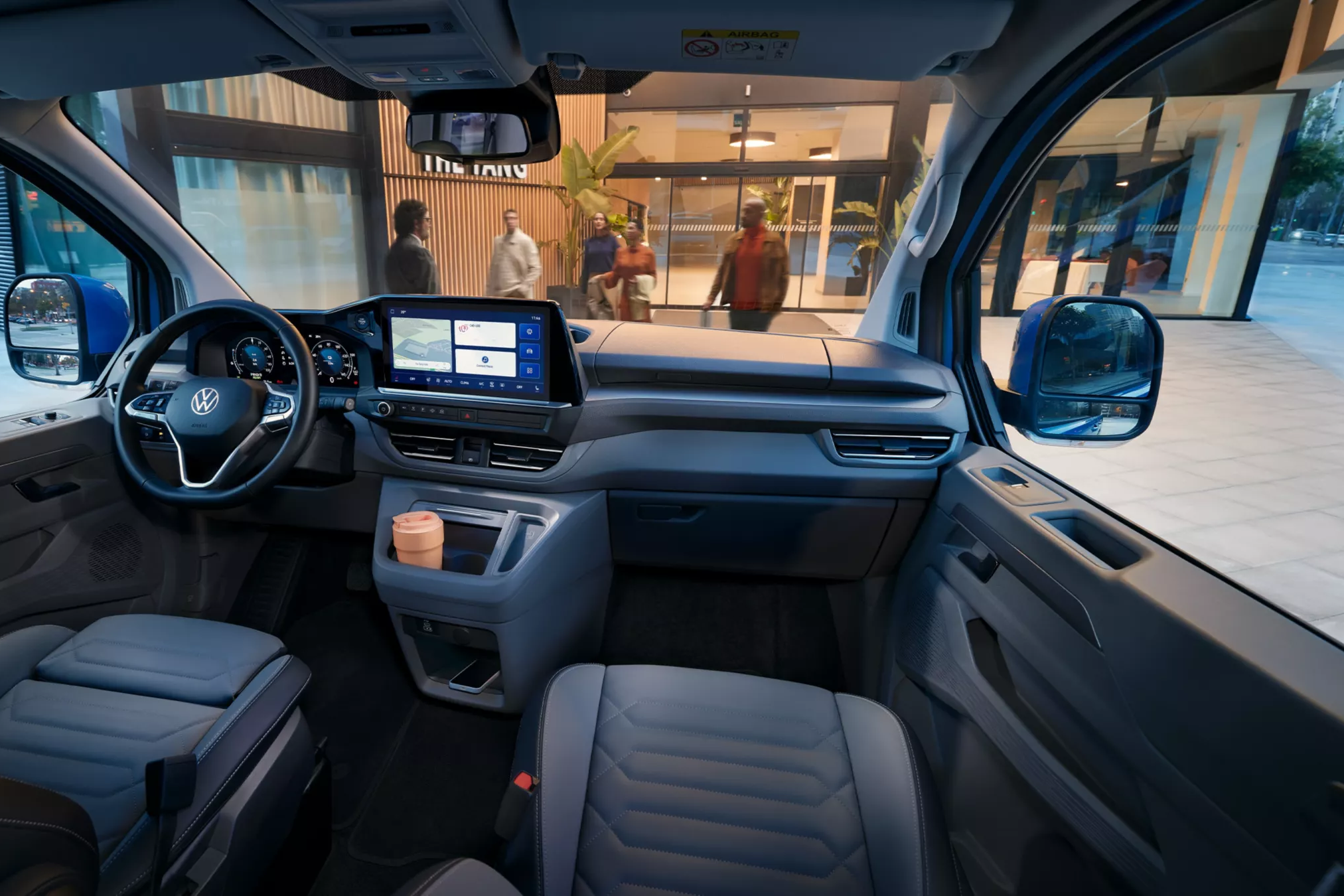 Interiér moderného automobilu značky Volkswagen s digitálnym prístrojovým panelom. Súčasťou sú dve predné sedadlá a držiak na nápoje s pohárom na kávu. Vozidlo je zaparkované pred nákupným centrom.