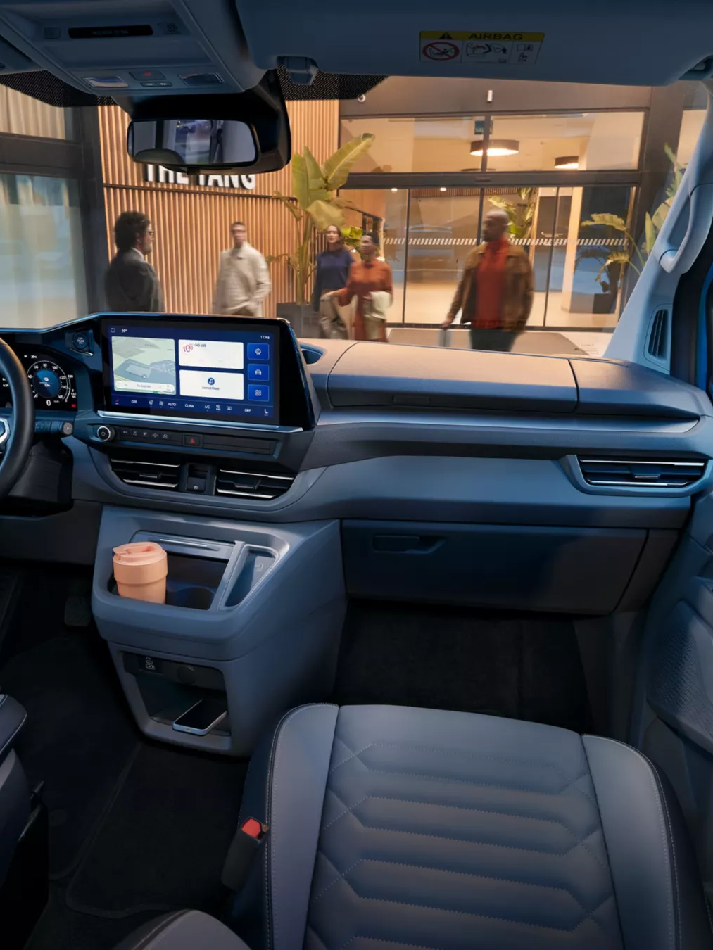 Interiér moderného automobilu značky Volkswagen s digitálnym prístrojovým panelom. Súčasťou sú dve predné sedadlá a držiak na nápoje s pohárom na kávu. Vozidlo je zaparkované pred nákupným centrom.