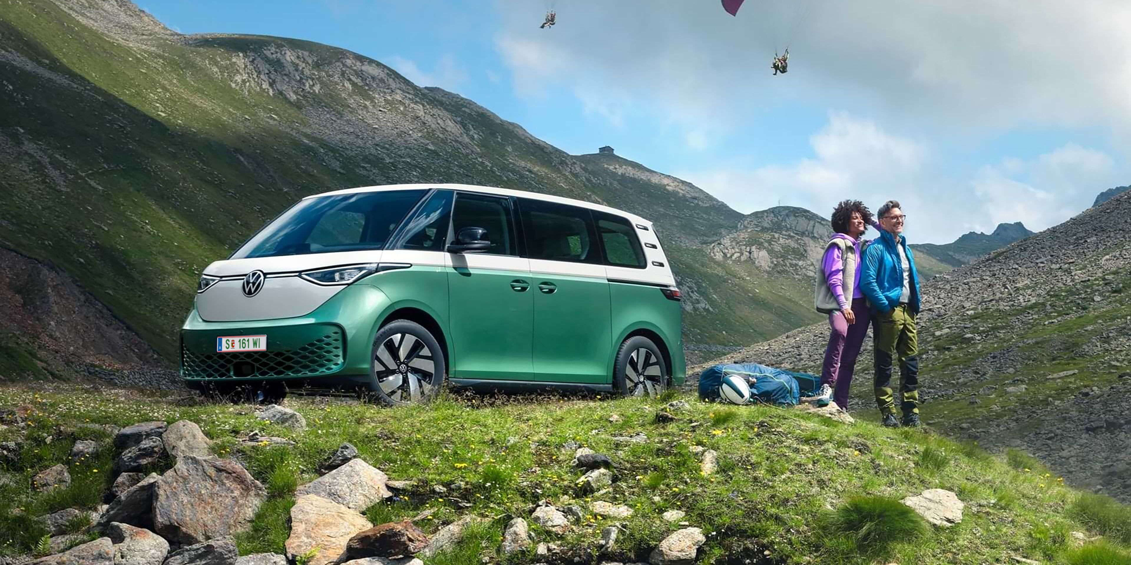 Dvojica stojí na zelenej lúke vedľa zeleného VW ID. Buzz. Na oblohe sú dvaja paraglidisti, ktorí lietajú nad kopcami. V pozadí vidieť kopce.