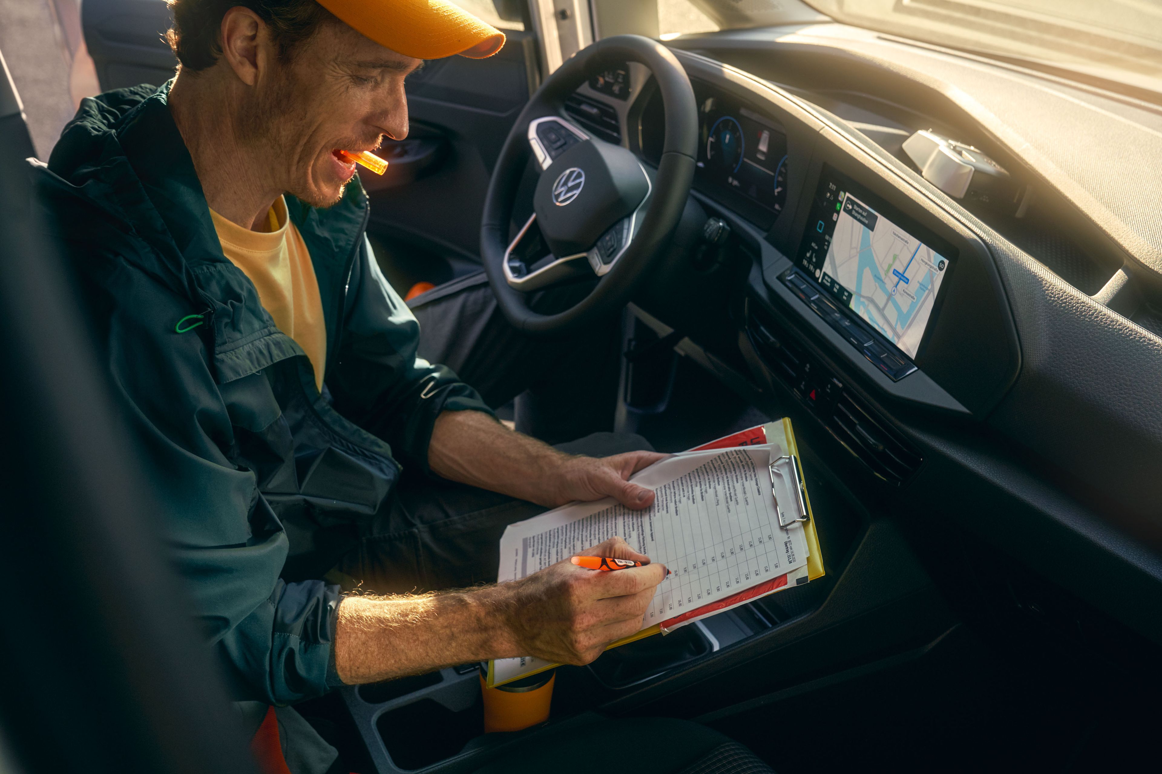 Muž sedí v aute a vypisuje údaje na papieri. Na dotykovom displeji systému infotainment je navigácia. Scéna je osvetlená žiarivým slnkom.