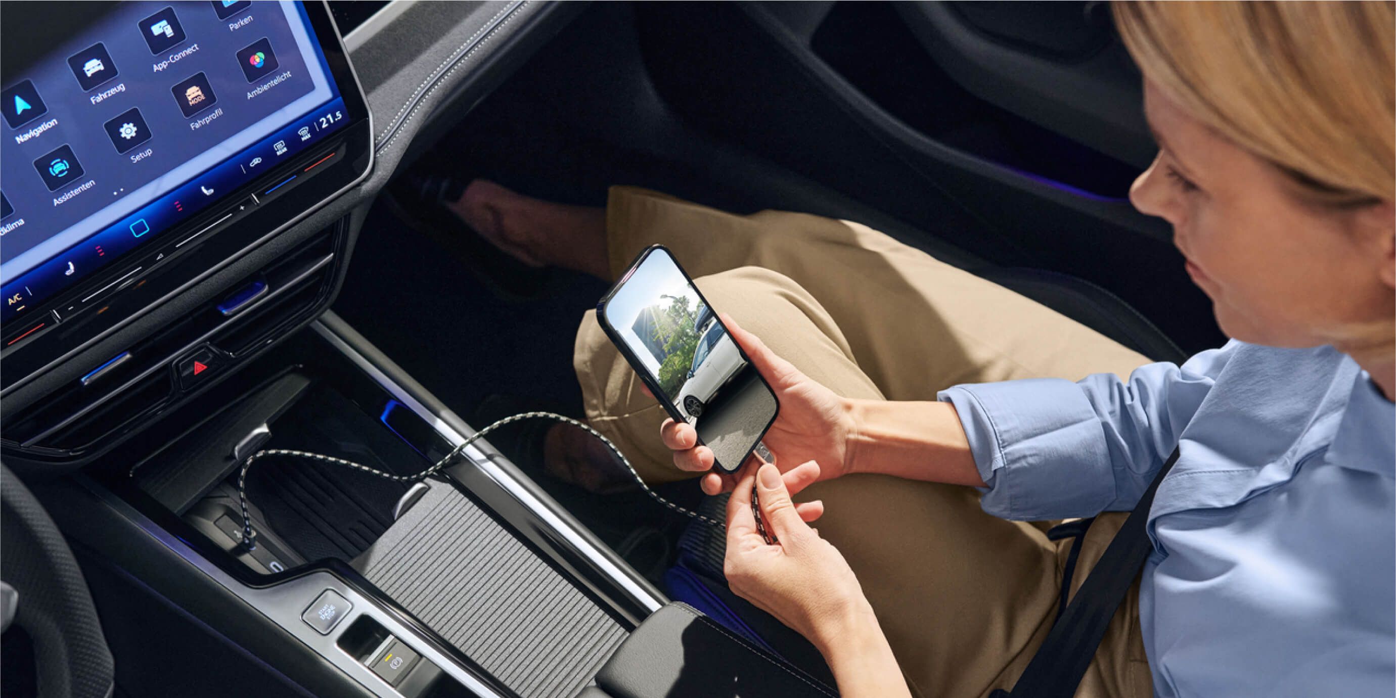 Žena sedí v aute a drží mobilný telefón pripojený USB káblom k vozidlu, aby sa nabíjal. Na displeji mobilu je vidieť cestu, ktorou prechádza auto. Na palubnej doske sú tiež viditeľné dotykové ovládacie panely a displej systému infotainment.