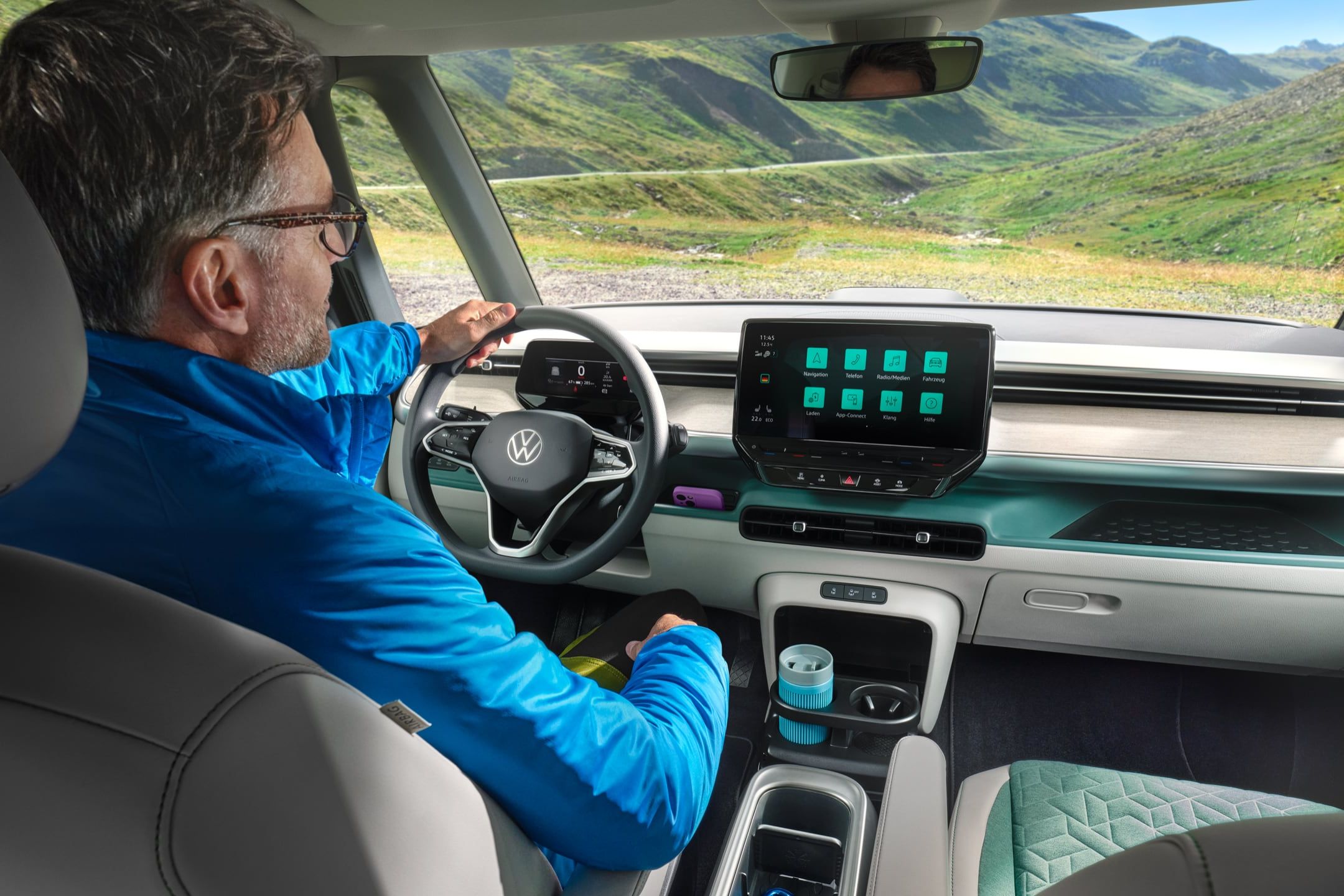 Muž sedí v modernom automobile s veľkým dotykovým displejom na palubnej doske. Cez čelné sklo vidieť horské prostredie a zelené údolie. Muž má modrú bundu.