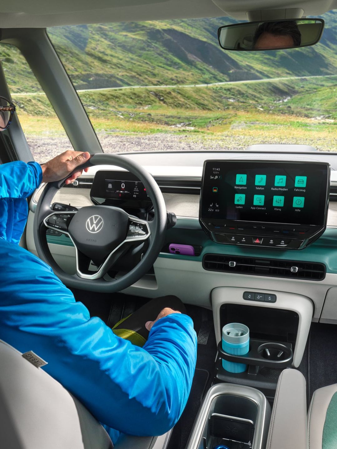 Muž sedí v modernom automobile s veľkým dotykovým displejom na palubnej doske. Cez čelné sklo vidieť horské prostredie a zelené údolie. Muž má modrú bundu.