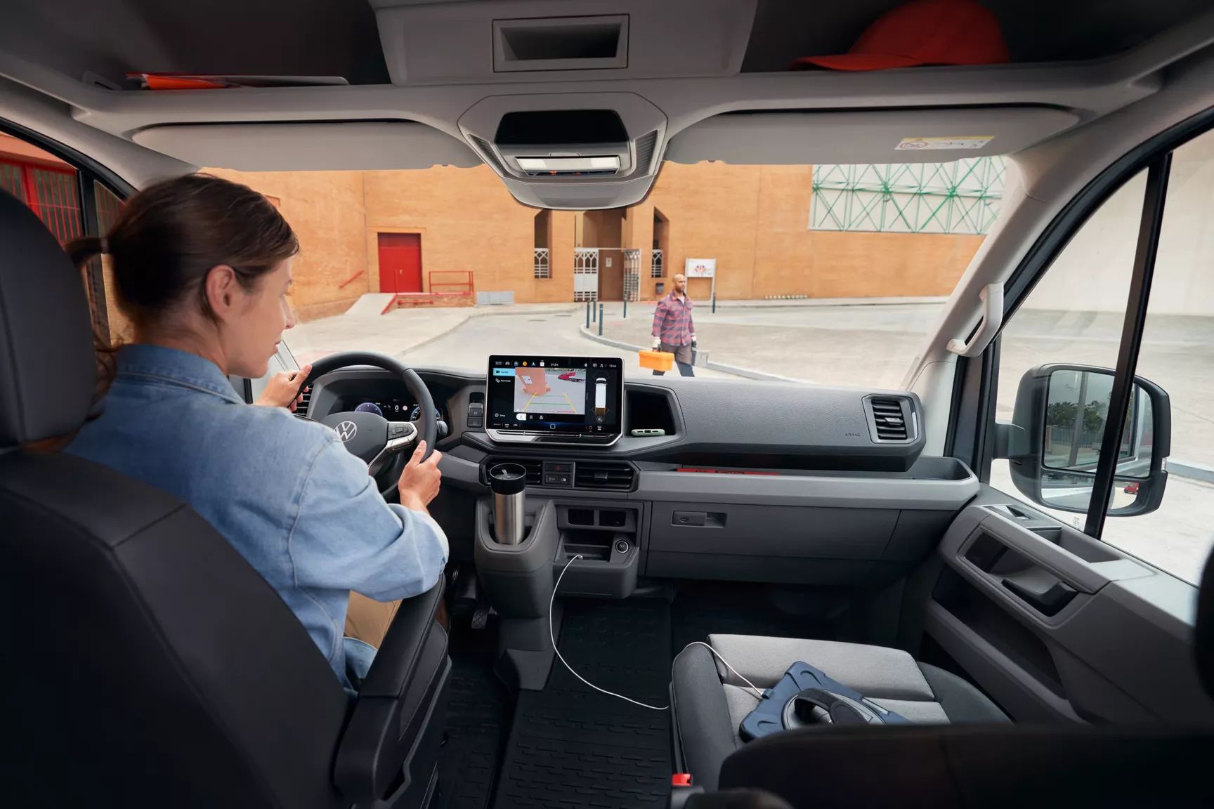 Žena sedí na sedadle vodiča vo vnútri vozidla a pozerá sa na displej systému infotainment s navigačným systémom. Muž kráča po ceste okolo vozidla. V interiéri auta vidieť niektoré prístroje a zariadenia na palubnej doske.
