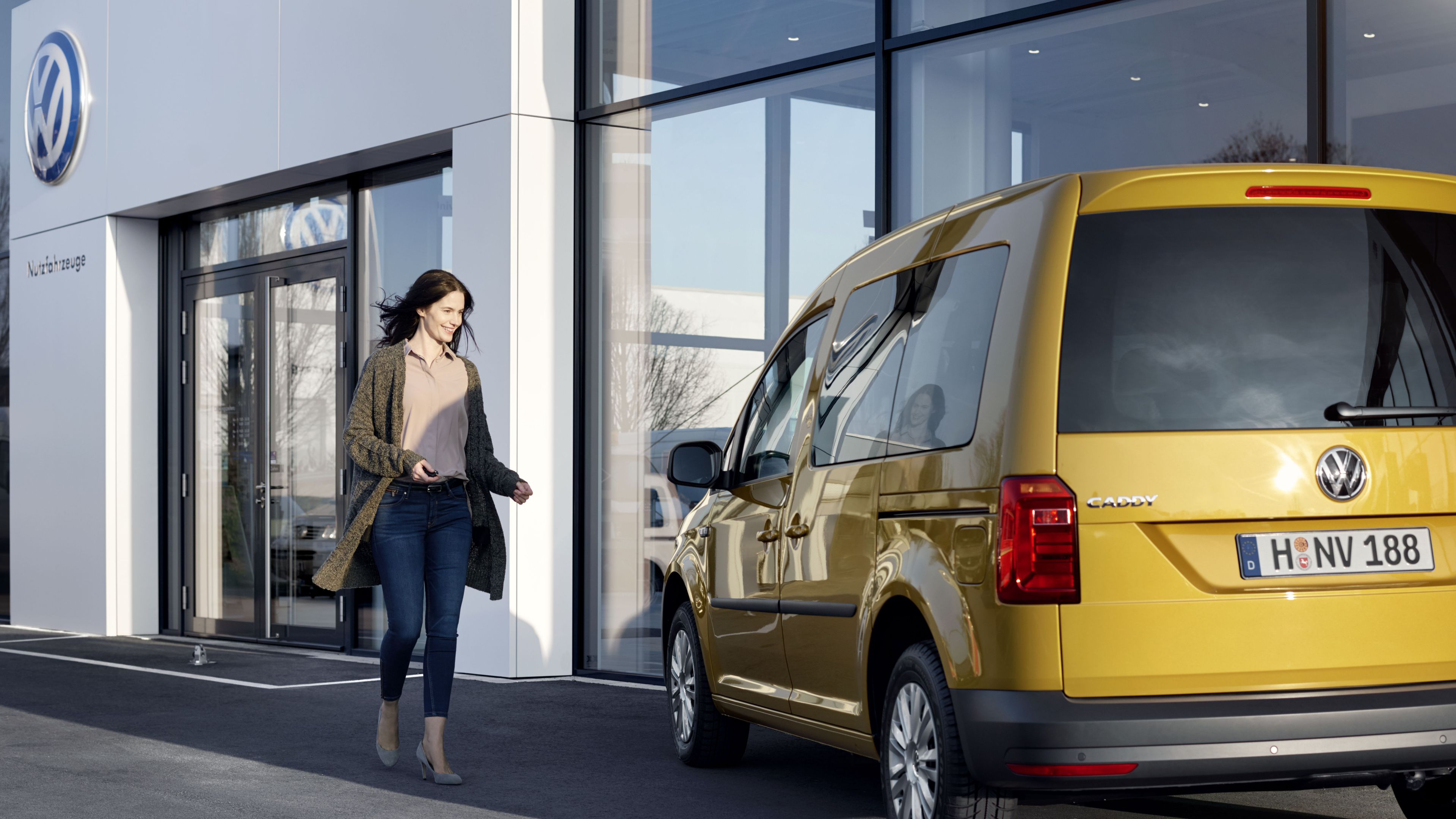 Žena kráča k žltému vozidlu Volkswagen Caddy, ktoré je zaparkované pred modernou budovou predajcu Volkswagen.