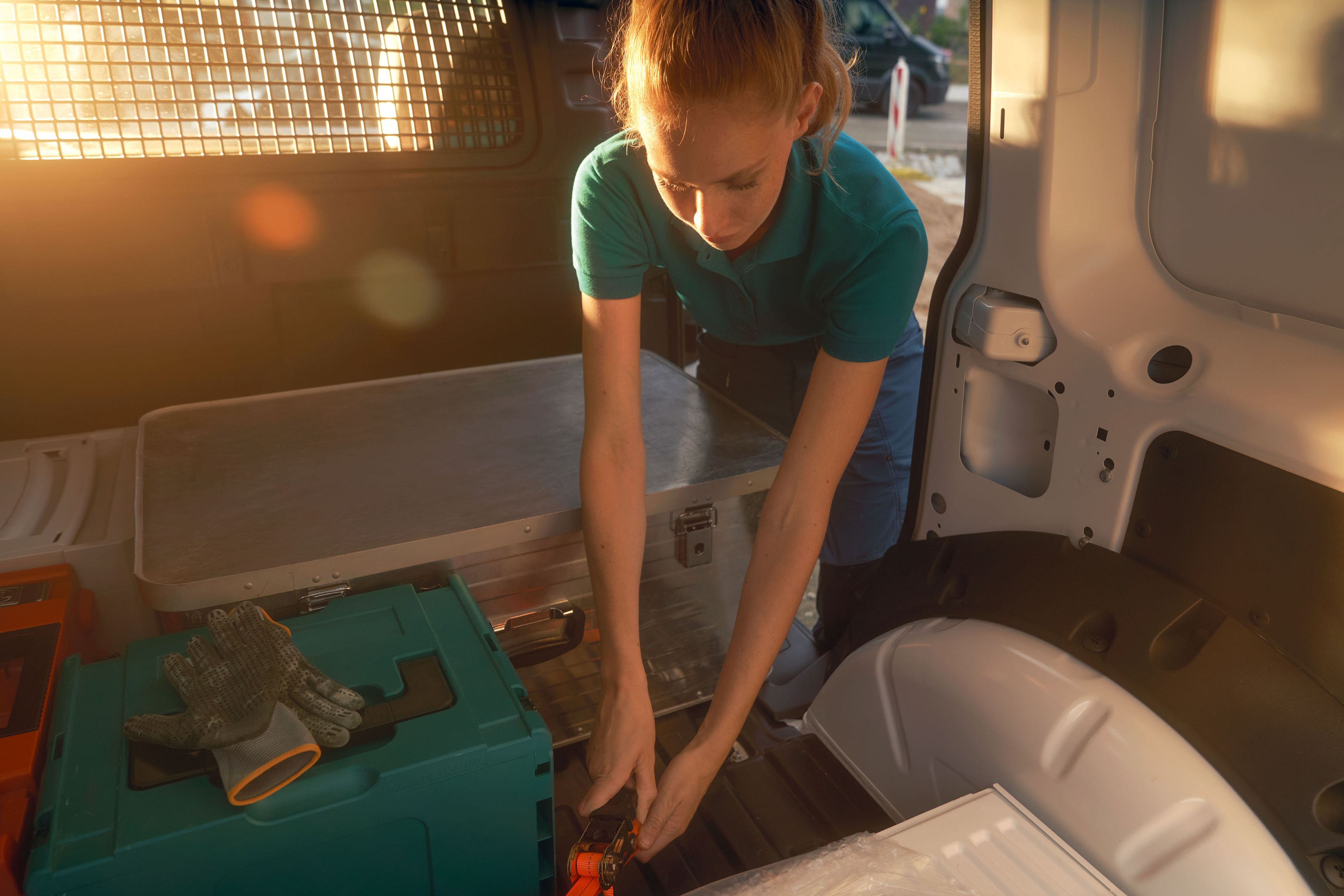 Žena v nákladnom priestore dodávky manipuluje s nástrojmi. Na pozadí je rozmazané slnko s oranžovými odleskami.