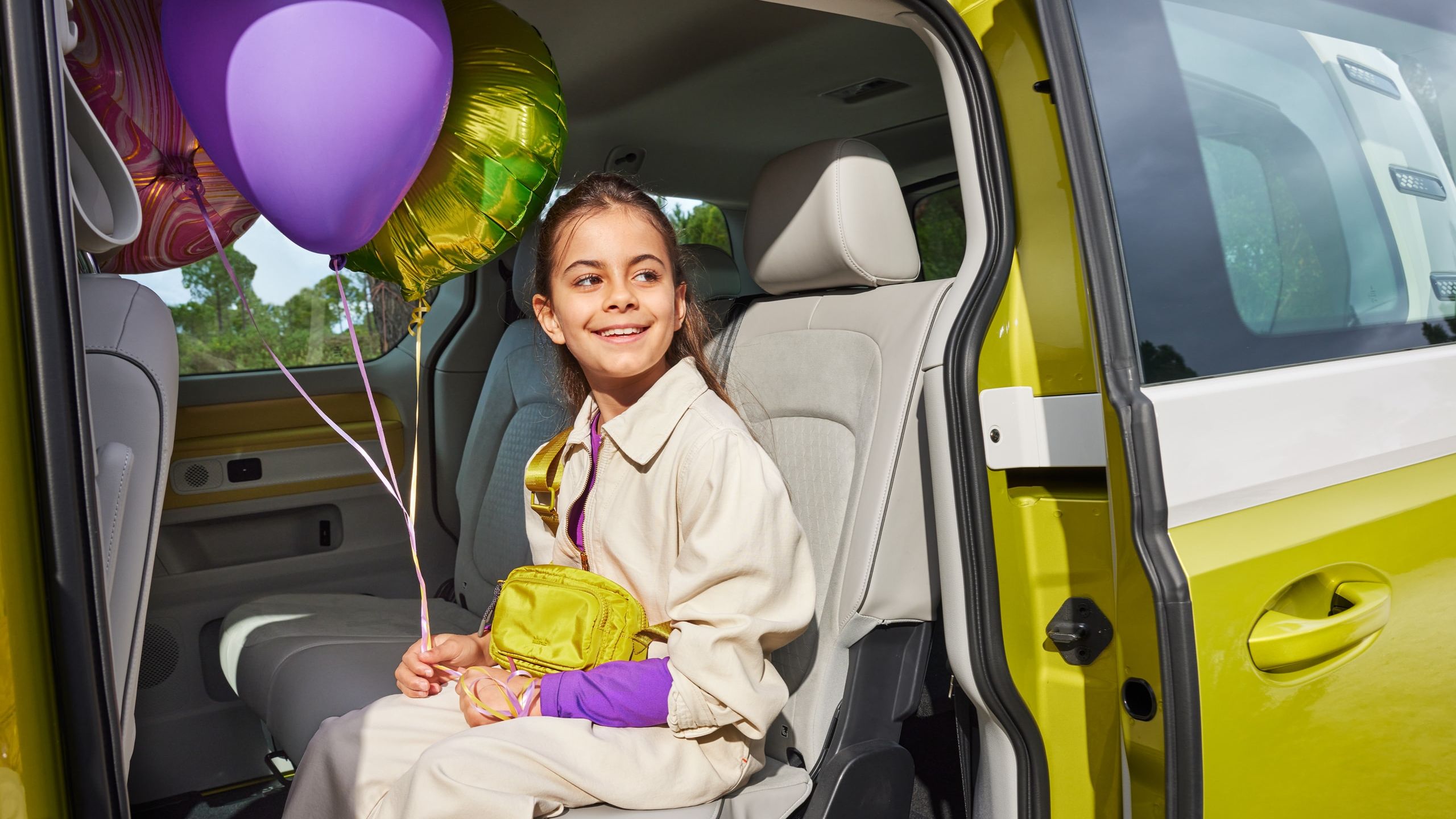 Lächelndes Kind mit Ballons sitzt auf Rücksitz eines gelb weißen ID. Buzz mit offener Schiebetür