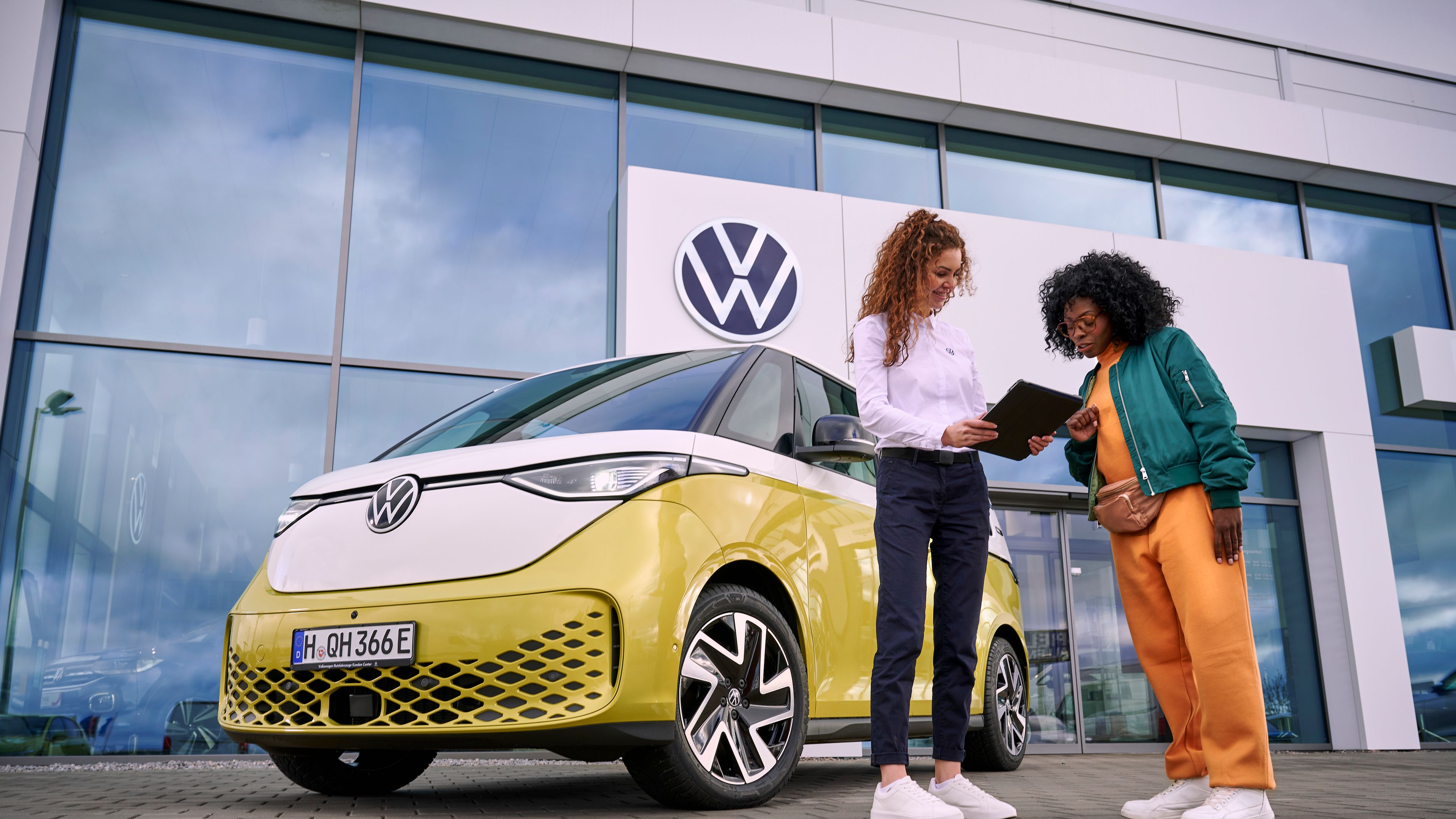 Dve ženy stoja pred novým žltým VW ID. Buzz pred predajňou. Jedna drží tablet a niečo na ňom ukazuje druhej žene. Obchodné stretnutie sa odohráva na parkovisku pred predajňou.
