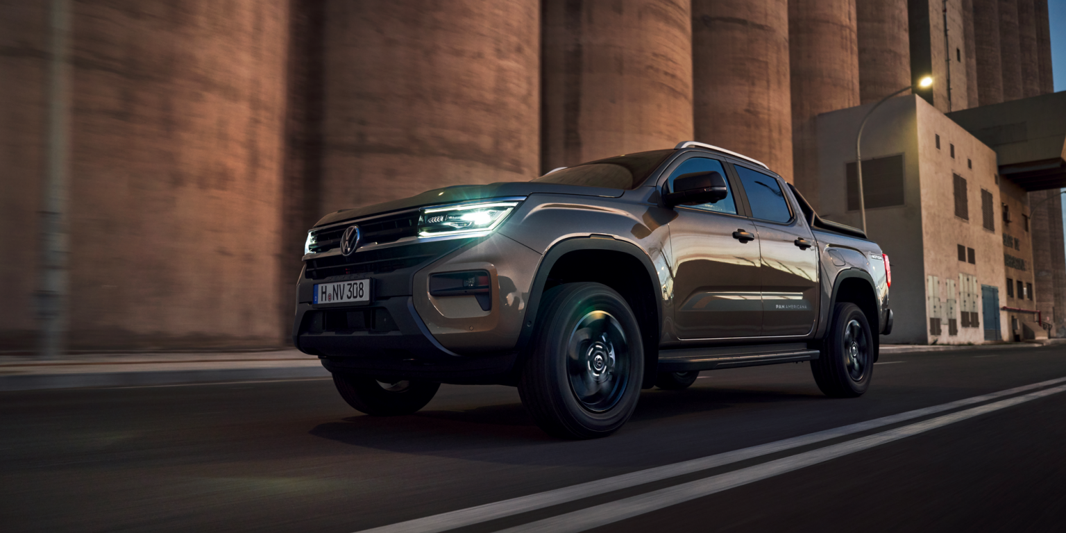 Tmavohnedý pickup Volkswagen jazdí po ceste pred veľkými priemyselnými budovami. Svetlá automobilu sú zapnuté a okolie je mierne osvetlené.
