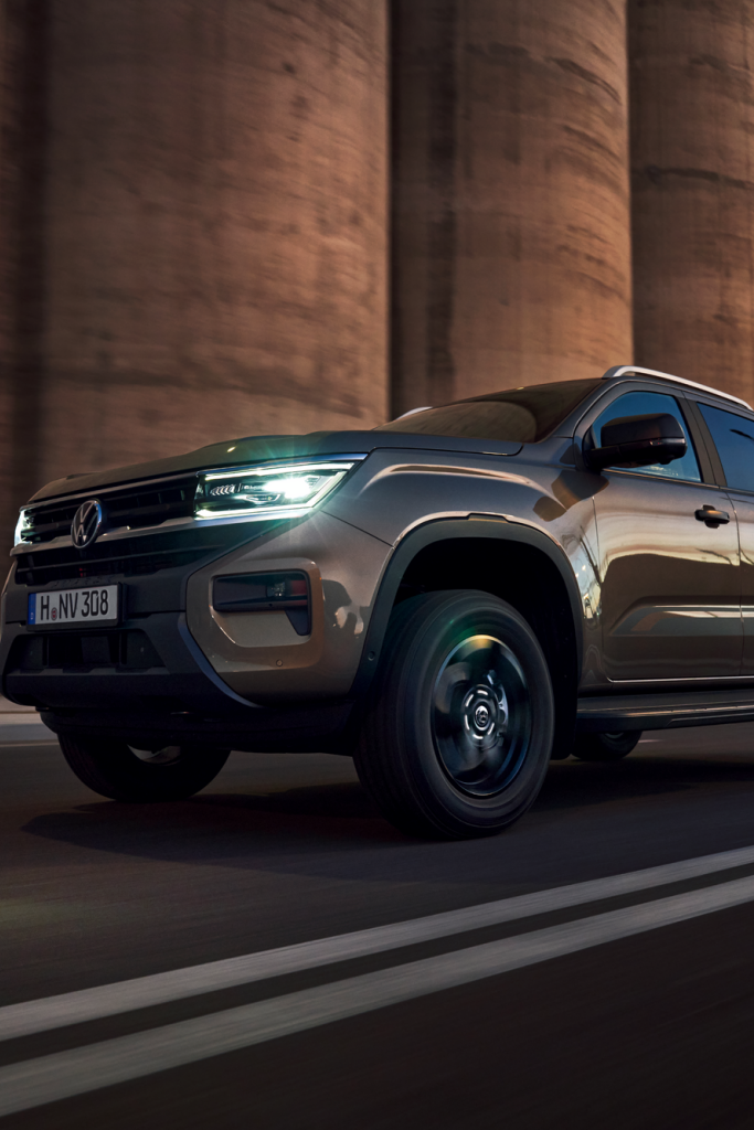 Tmavohnedý pickup Volkswagen jazdí po ceste pred veľkými priemyselnými budovami. Svetlá automobilu sú zapnuté a okolie je mierne osvetlené.