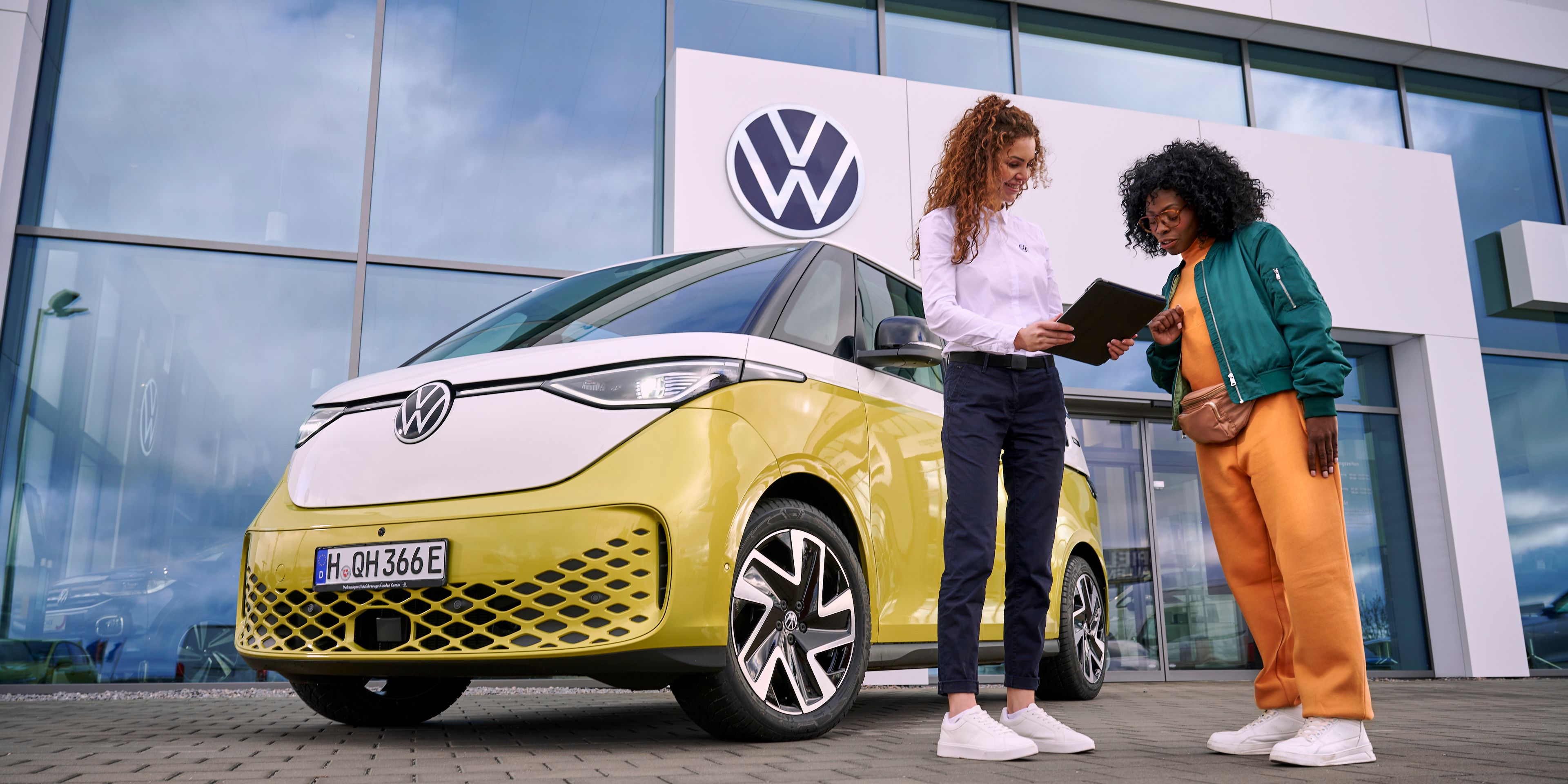 Dve ženy stoja pred novým žltým VW ID. Buzz pred predajňou. Jedna drží tablet a niečo na ňom ukazuje druhej žene. Obchodné stretnutie sa odohráva na parkovisku pred predajňou.