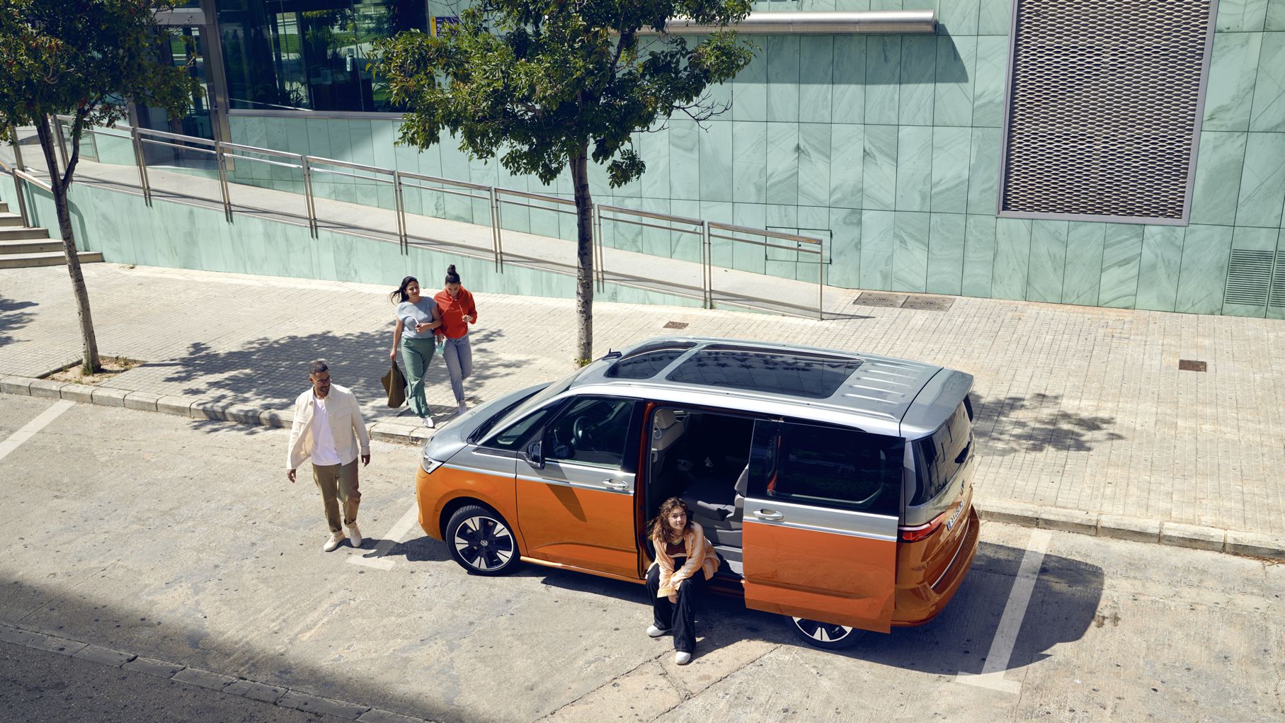 VW Multivan Energetic stojí na parkovacom mieste pri chodníku. Bočné dvere sú otvorené a vo vozidle sedí mlada žena. K automobilu prichádzajú traja ďalší ľudia. Za vozidlom je budova so svetlozelenou fasádou.