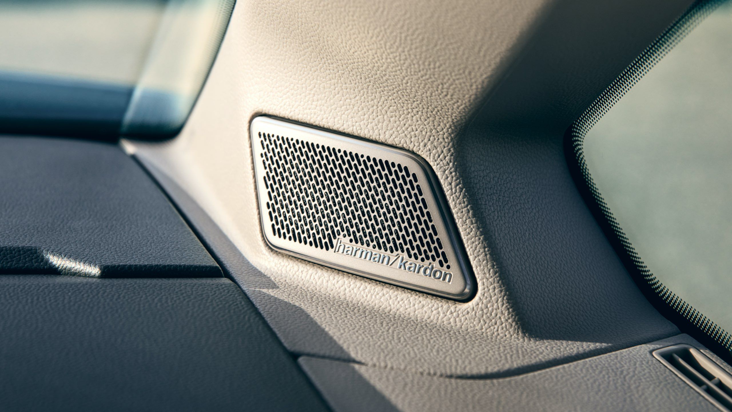 Detailný záber na audiosystém Harman Kardon vo vnútri automobilu. Reproduktor je integrovaný do plastového panelu pri prednom okne. Na mriežke je názov značky "Harman Kardon".