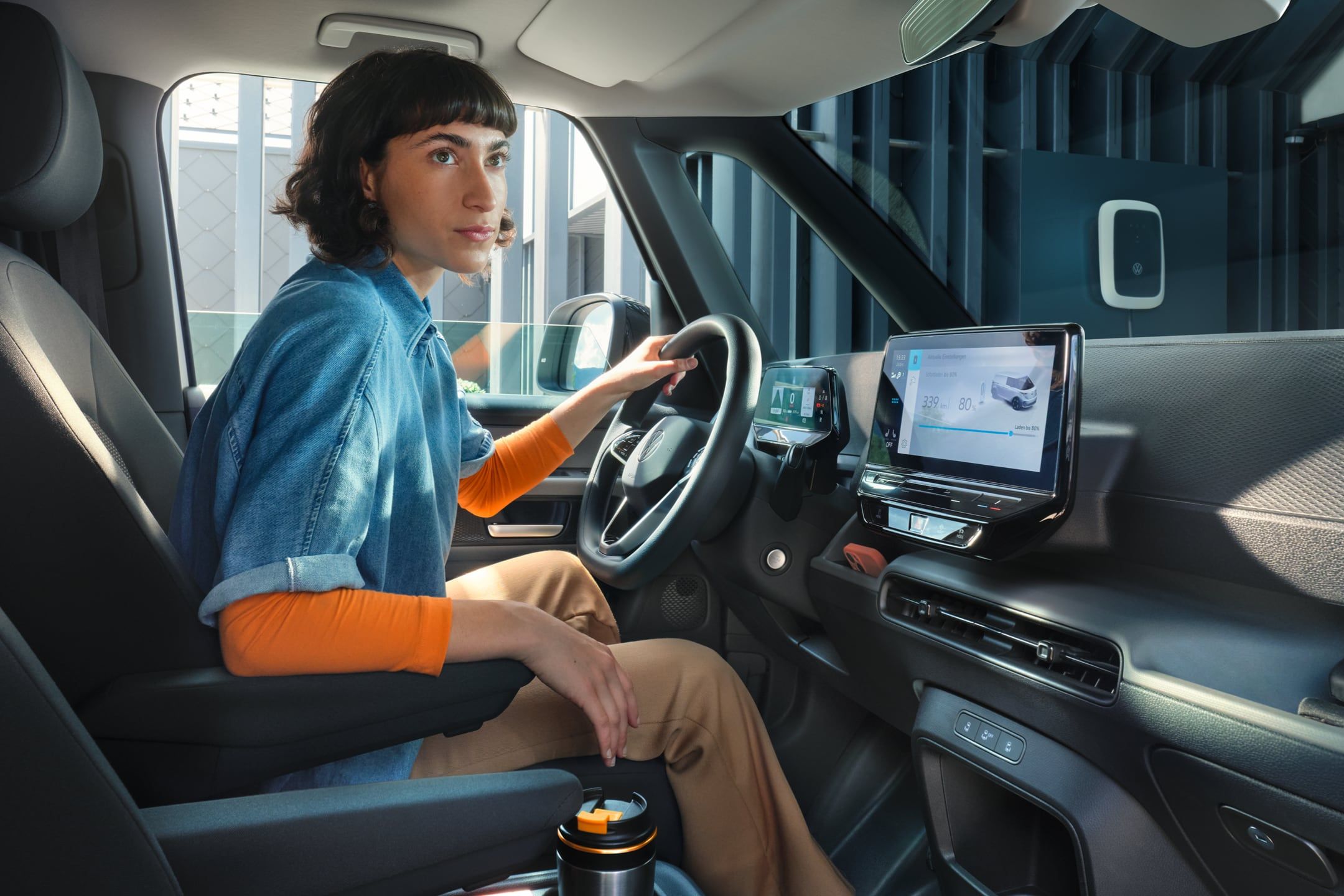 Žena sedí za volantom auta, ktoré má moderný interiér vybavený dotykovým displejom systému infotainment na palubnej doske. Cez čelné a bočné okno vidieť nabíjaciu stanicu pre elektrické vozidlá. 