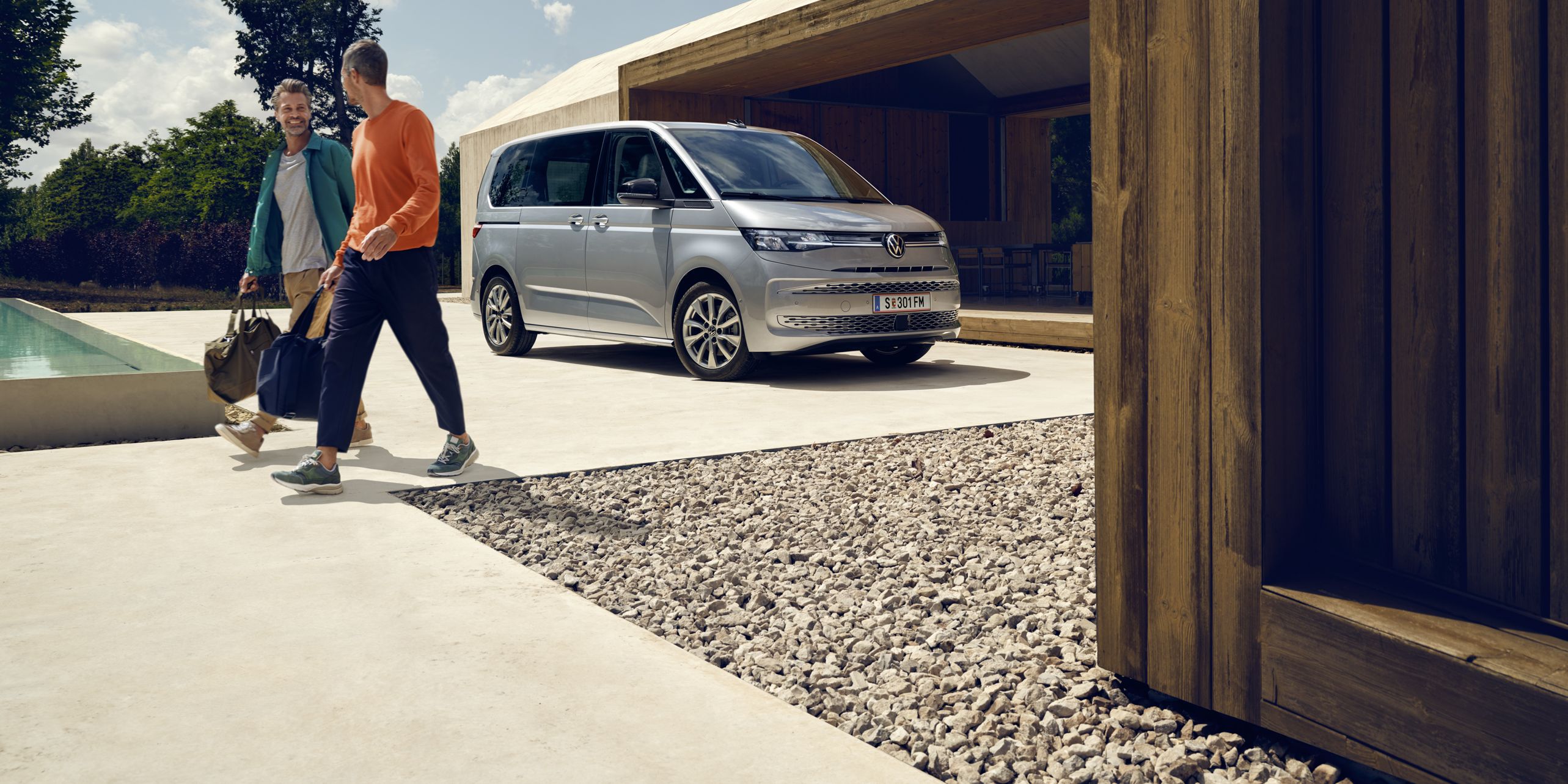 Dvaja muži kráčajú popri bazéne pri modernej vile. Pri dome je zaparkovaný strieborný VW Multivan. Okolité stromy a modrá obloha vytvárajú príjemnú scénu.