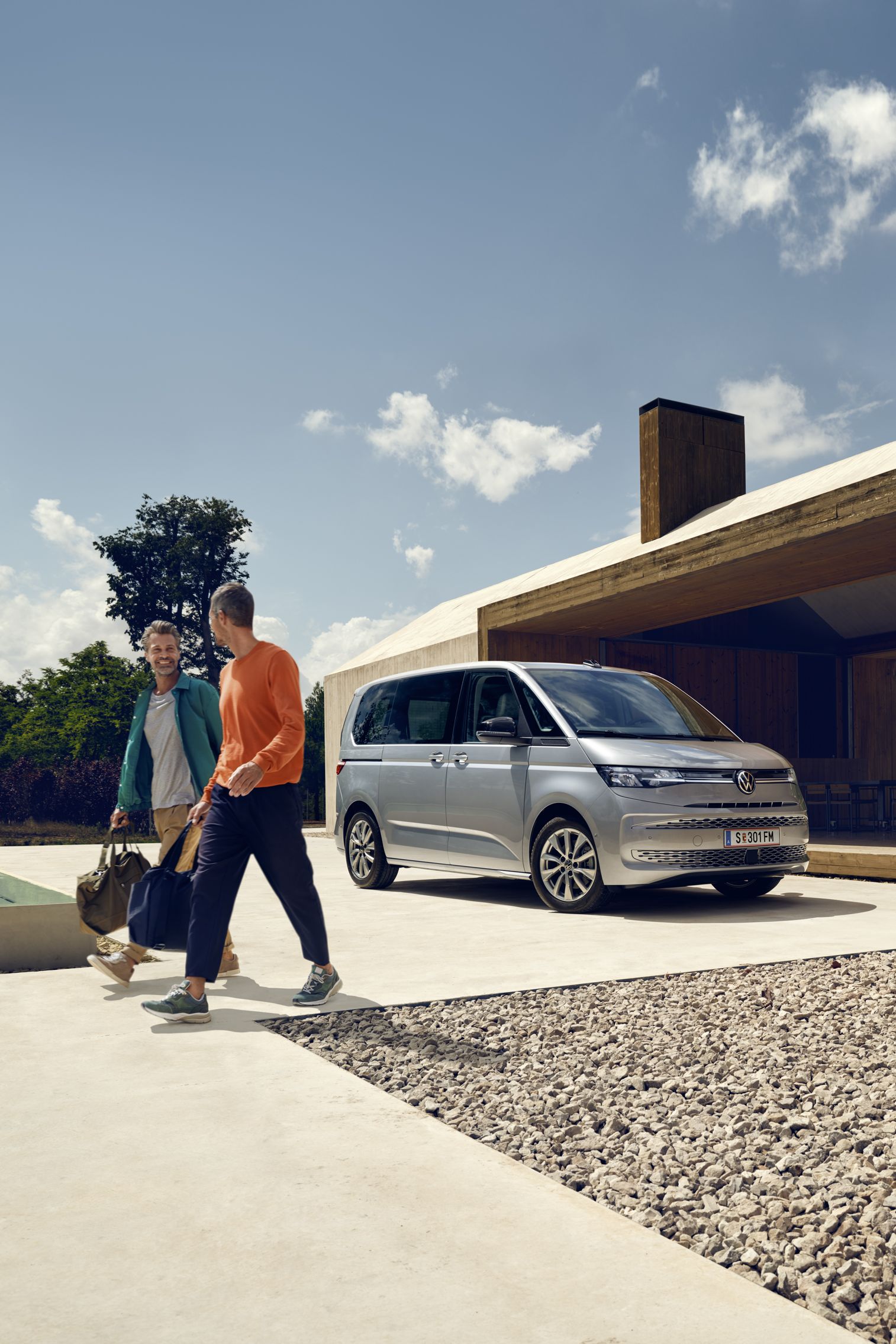Dvaja muži kráčajú popri bazéne pri modernej vile. Pri dome je zaparkovaný strieborný VW Multivan. Okolité stromy a modrá obloha vytvárajú príjemnú scénu.