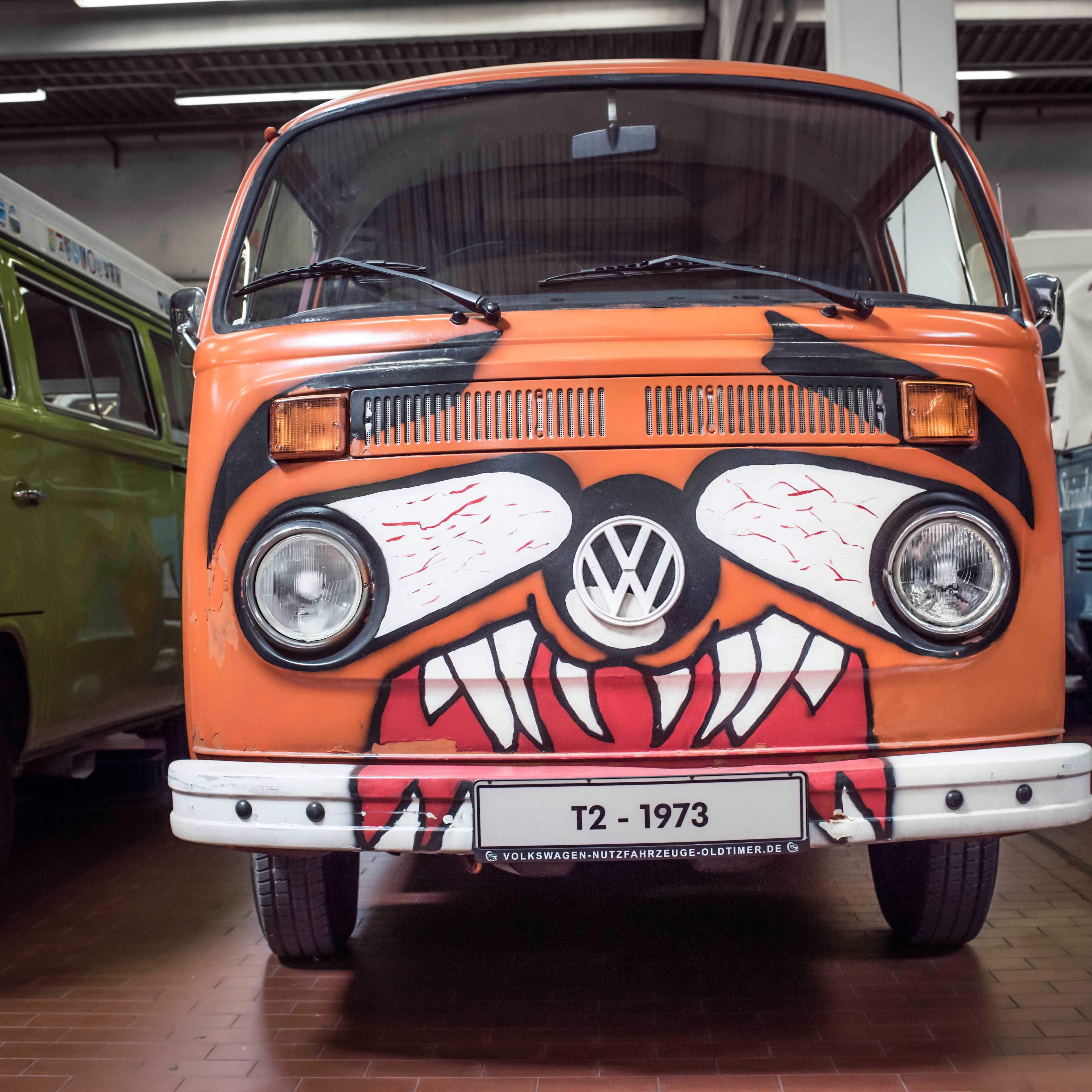 Oranžová dodávka VW T2 z roku 1973 s predným nárazníkom upraveným do podoby tváre draka. Okolo sú ďalšie staré dodávky. Automobil je vystavený v krytom priestore.