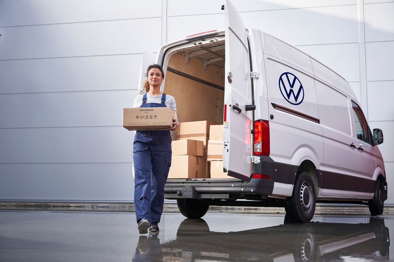 Žena v pracovnom oblečení nesie kartónovú krabicu z otvoreného nákladného vozidla značky Volkswagen. V pozadí je stena budovy. Vozidlo a žena sa odrážajú v mokrej ceste..