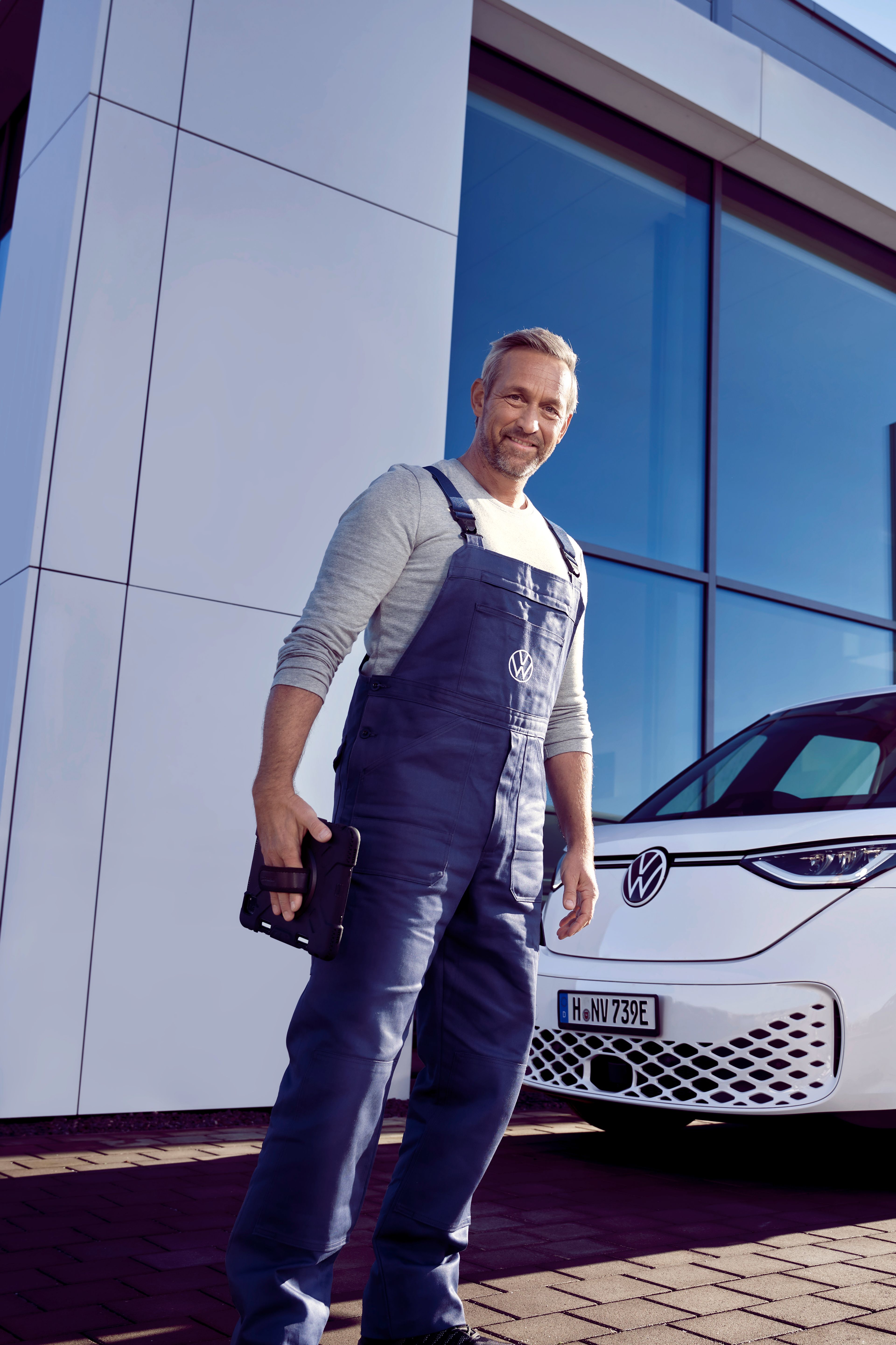 Servisný pracovník v modrých montérkach stojí pred bielym ID. Buzz, ktorí je zaparkovaný pred showroomom Volkswagen. Muž sa pozerá priamo do objektívu.