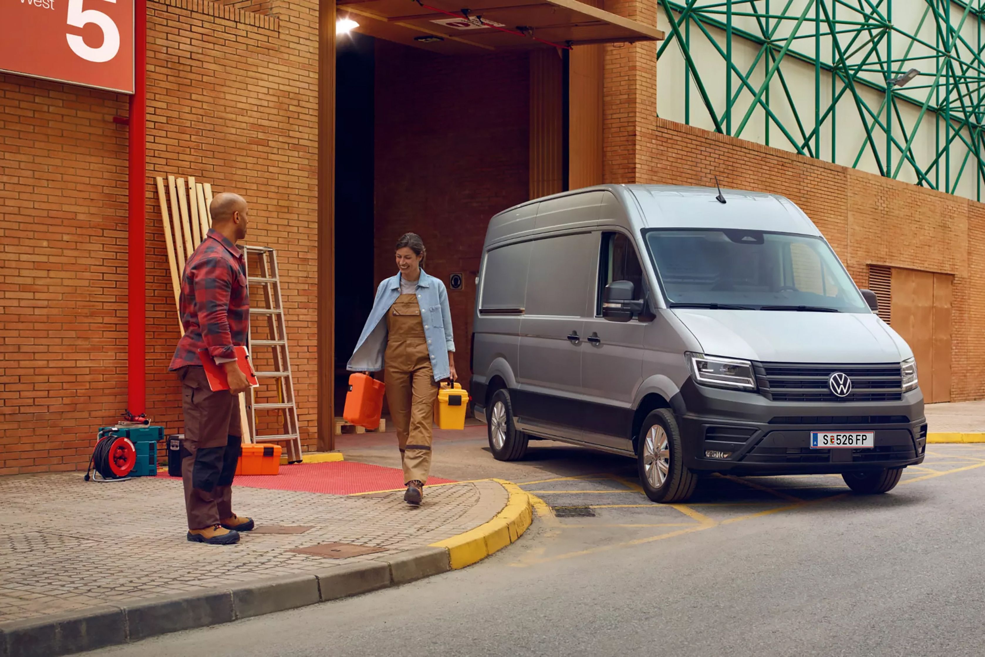 Dvaja pracovníci, muž a žena, sú pred vchodom do budovy a žena nesie nástroje muž stojí na chodníku s formulárom a pozerá sa na ňu. Vedľa nich je zaparkovaný Crafter dodávka. Pozadie tvorí tehlová budova s kovovou konštrukciou.