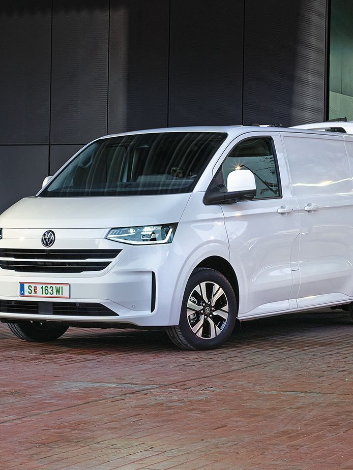 Biely e-Transporter zaparkovaný pred modernou budovou s veľkými oknami a modernou architektúrou. Pri budove je verejná nabíjacia stanica, na ktorej sa vozidlo nabíja. Za dodávkou sa nachádzajú dve osoby pri drevených debn8ch. Okolie je vydláždené červenými tehlovými kockami.